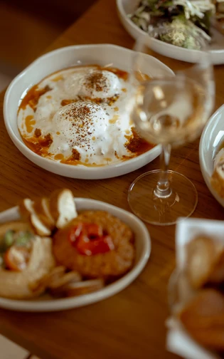 A wooden table with dishes of poached eggs in tomato sauce, salad, bread, dips, and wine.