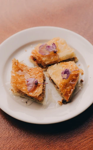 Three pieces of baklava on a white plate, drizzled with honey and garnished with purple flowers
