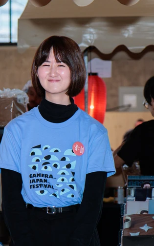 A CAMERA JAPAN team member stands at a festival stall beside a red lantern.
