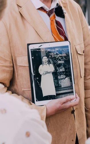 een hand houd een ipad vast waarop een zwart wit foto te zien is, met een man voor een gevel.