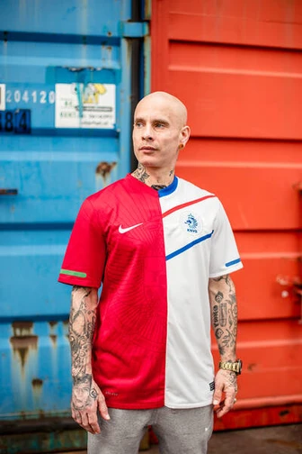 A tattooed man in a red-and-white Netherlands football shirt stands in front of colourful shipping c