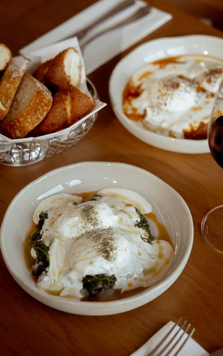 Two plates of poached eggs with yogurt and herbs sit on a wooden table,