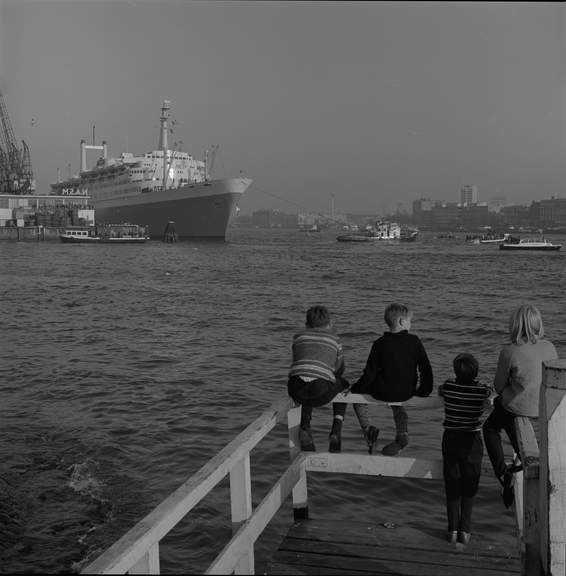 Jongens zitten op een Katendrechtse steiger en kijken richting SS Rotterdam, afgemeerd aan de Wilhelminakade