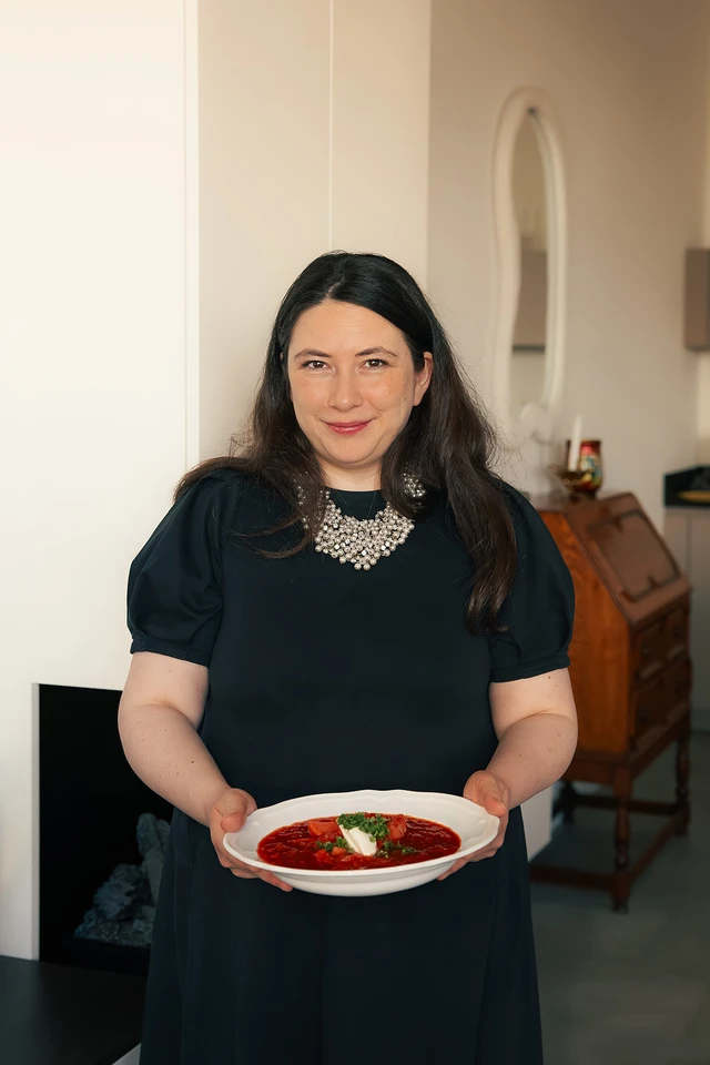 Woman in a dark dress, holding a plate of borscht with cream in a warmly lit room.