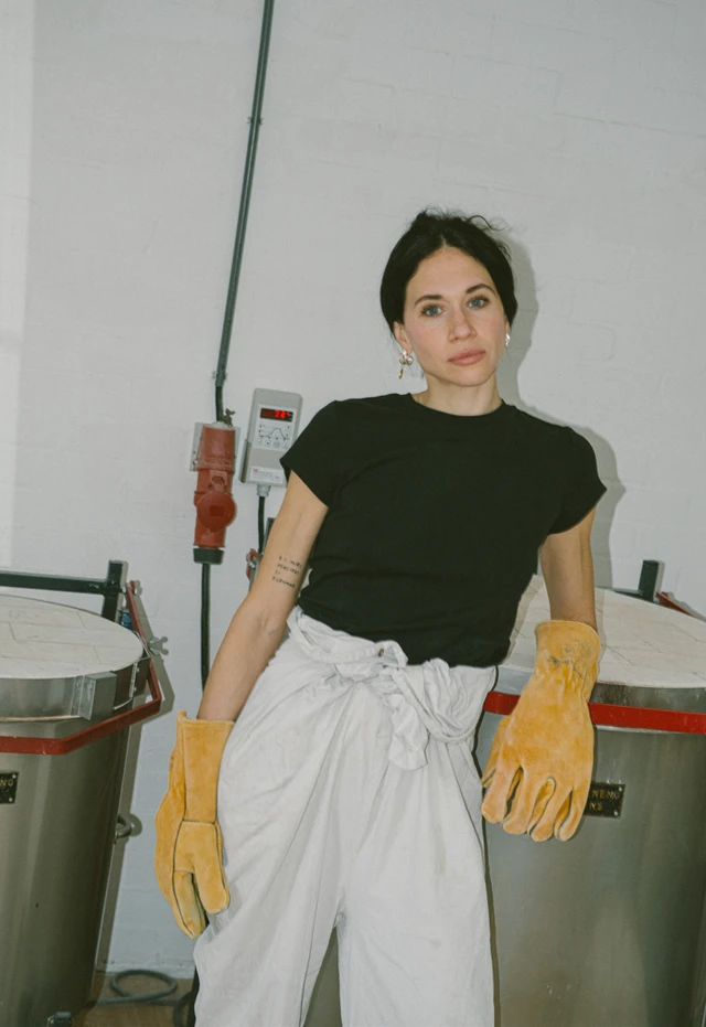 A ceramic artist wearing protective gloves stands between two large kilns in her studio.