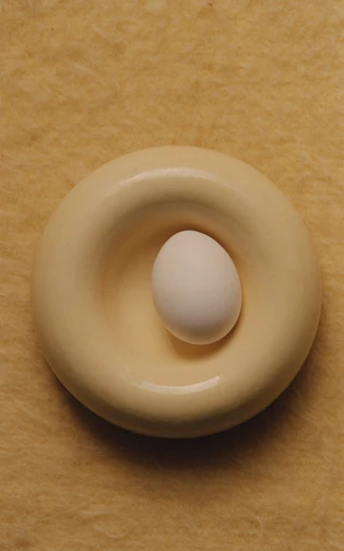 A white egg resting in a rounded yellow ceramic dish on a textured ochre surface.