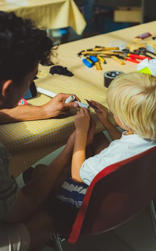 An adult helps a child make an object from white tubes at a craft table.