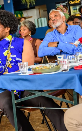 Visitors sit together at tables, listening to the programme during a Cabo Verdean cultural gathering