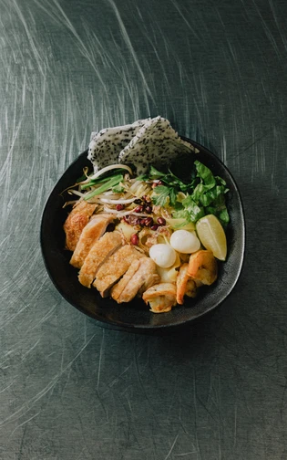 A black bowl holds: noodles, grilled chicken, prawns, leafy greens, lime wedges and quail eggs.