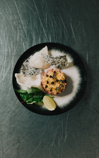 A black and white plate with a tartare, leafy greens, lime wedge, and speckled crisps.