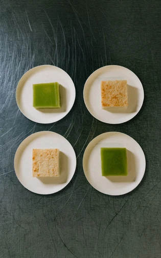 Four small plates on a metal surface, each with a green or beige dessert square.