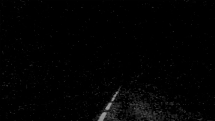A dark, grainy image of a road at night, with a faintly visible dashed white line