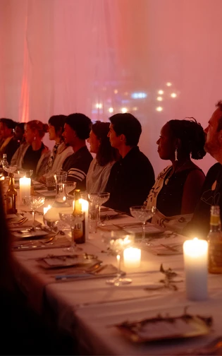 A diverse group of people sit at a long, elegantly set table, softly lit by candles.