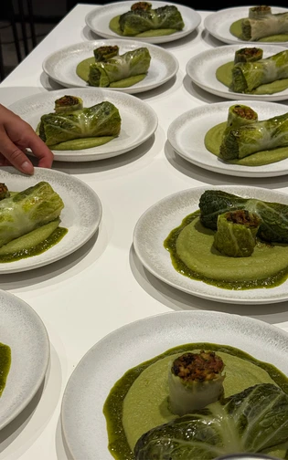 Rows of plates with stuffed cabbage rolls sit atop green puree. A hand arranges one plate.